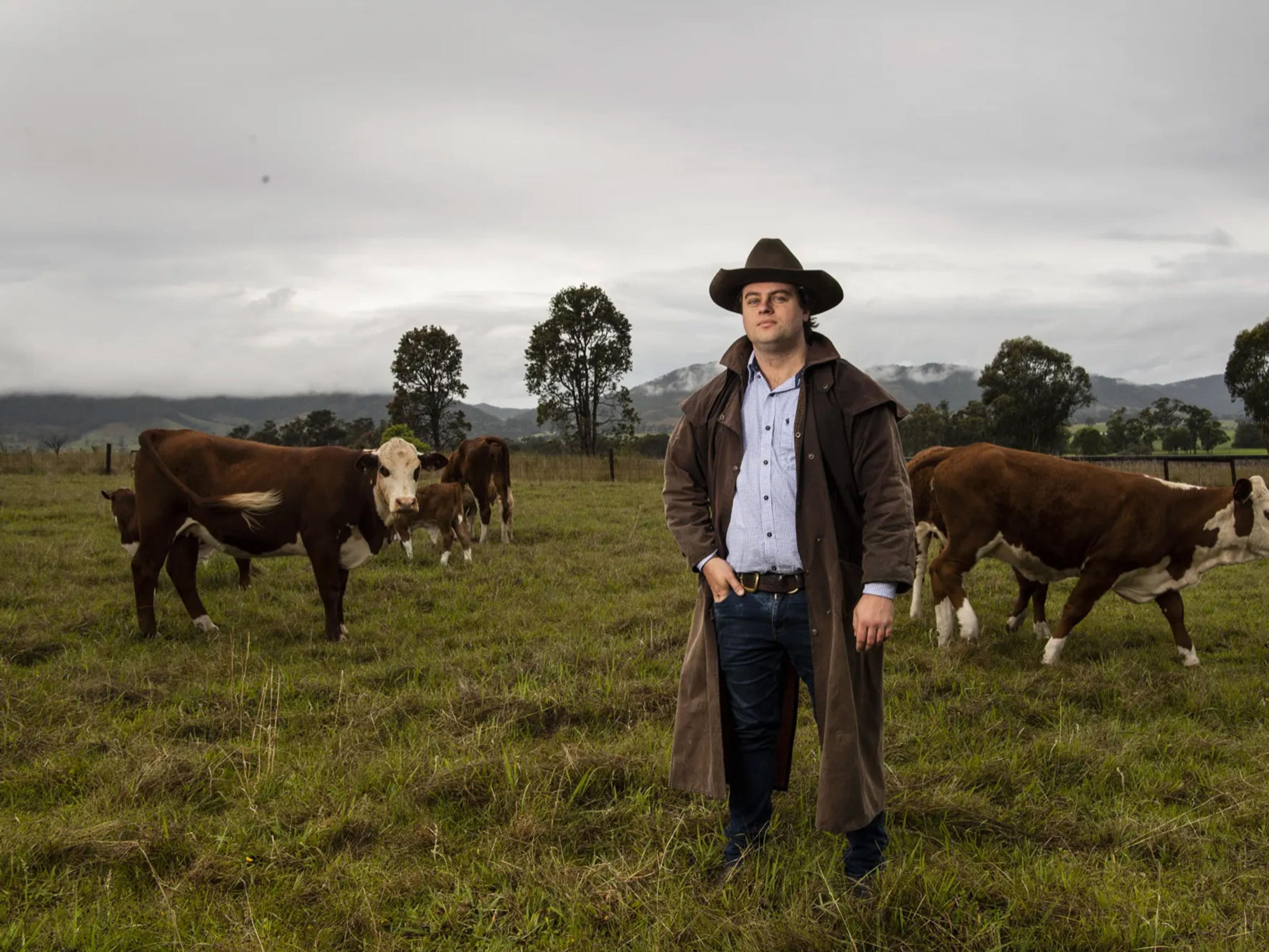A man starring into with a broad brimmed hat in a paddock with cows. The sky is cloudy.