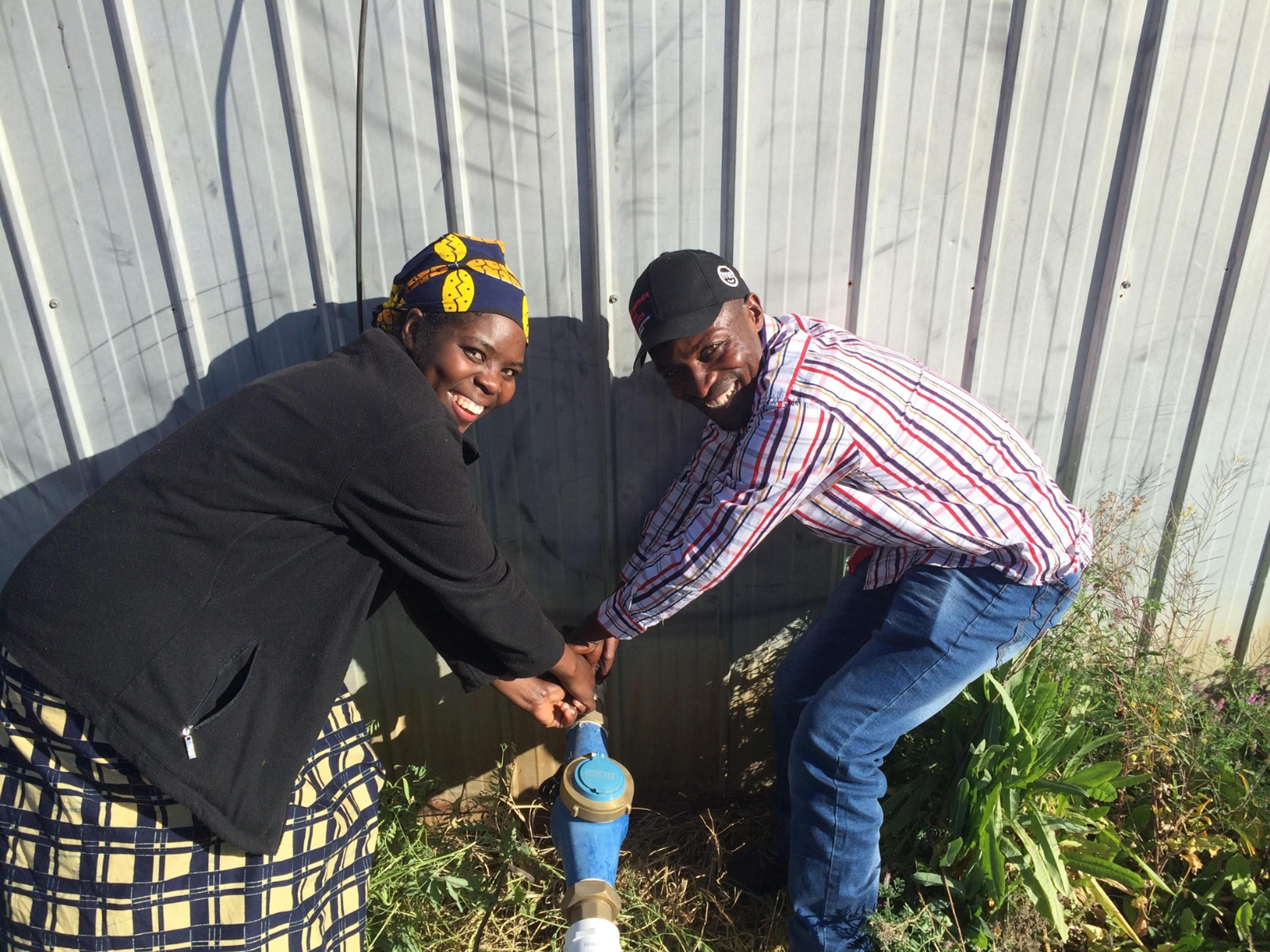 Two people turning on an outside tap and smiling at the camera.