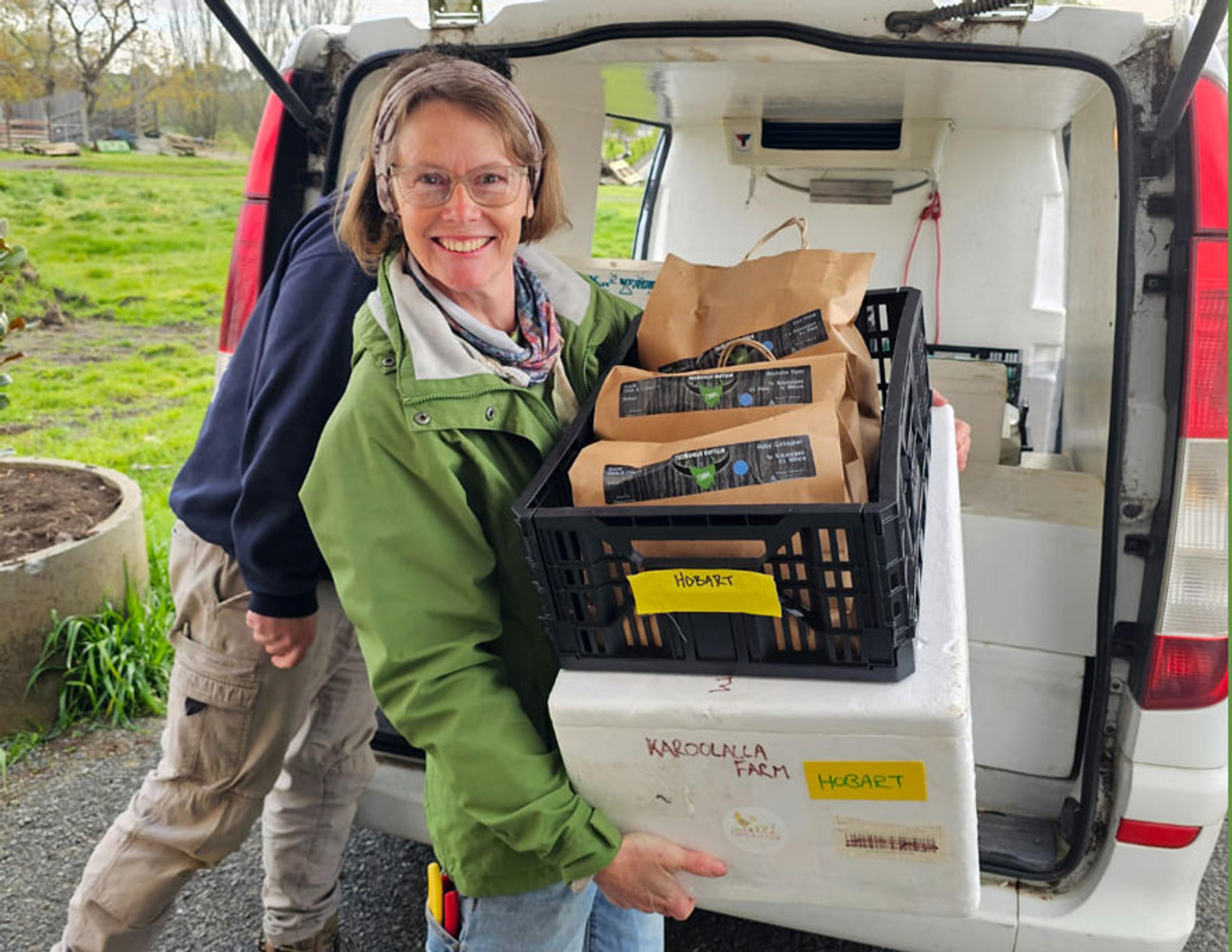 A smiling woman loading a crate and a cool box into a van. The crate has paper bags presumably filled with fresh food.