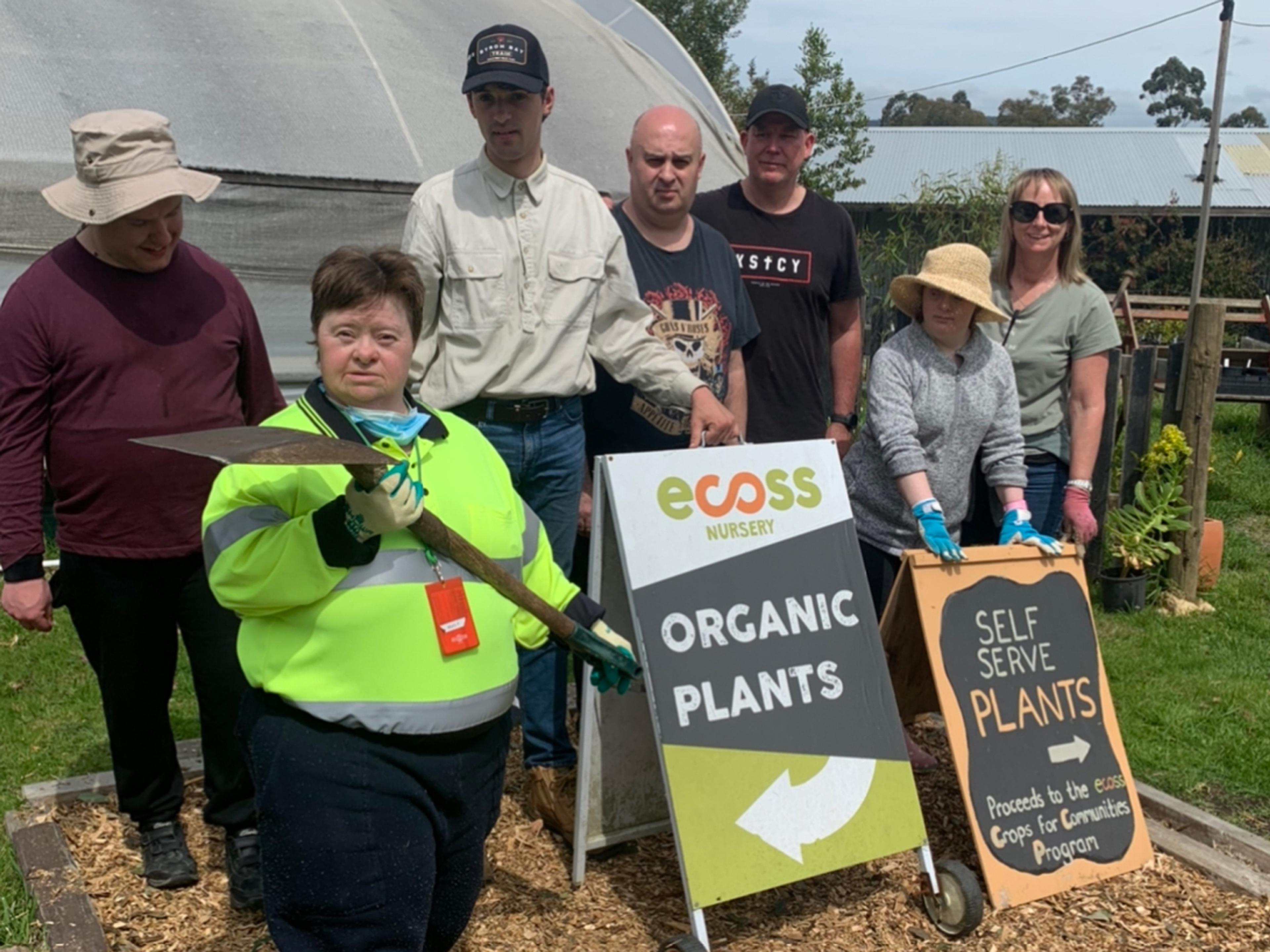 A group of people standing in front of a hoop house. They are standing behind two signs that indicate they are selling plants and seeds.