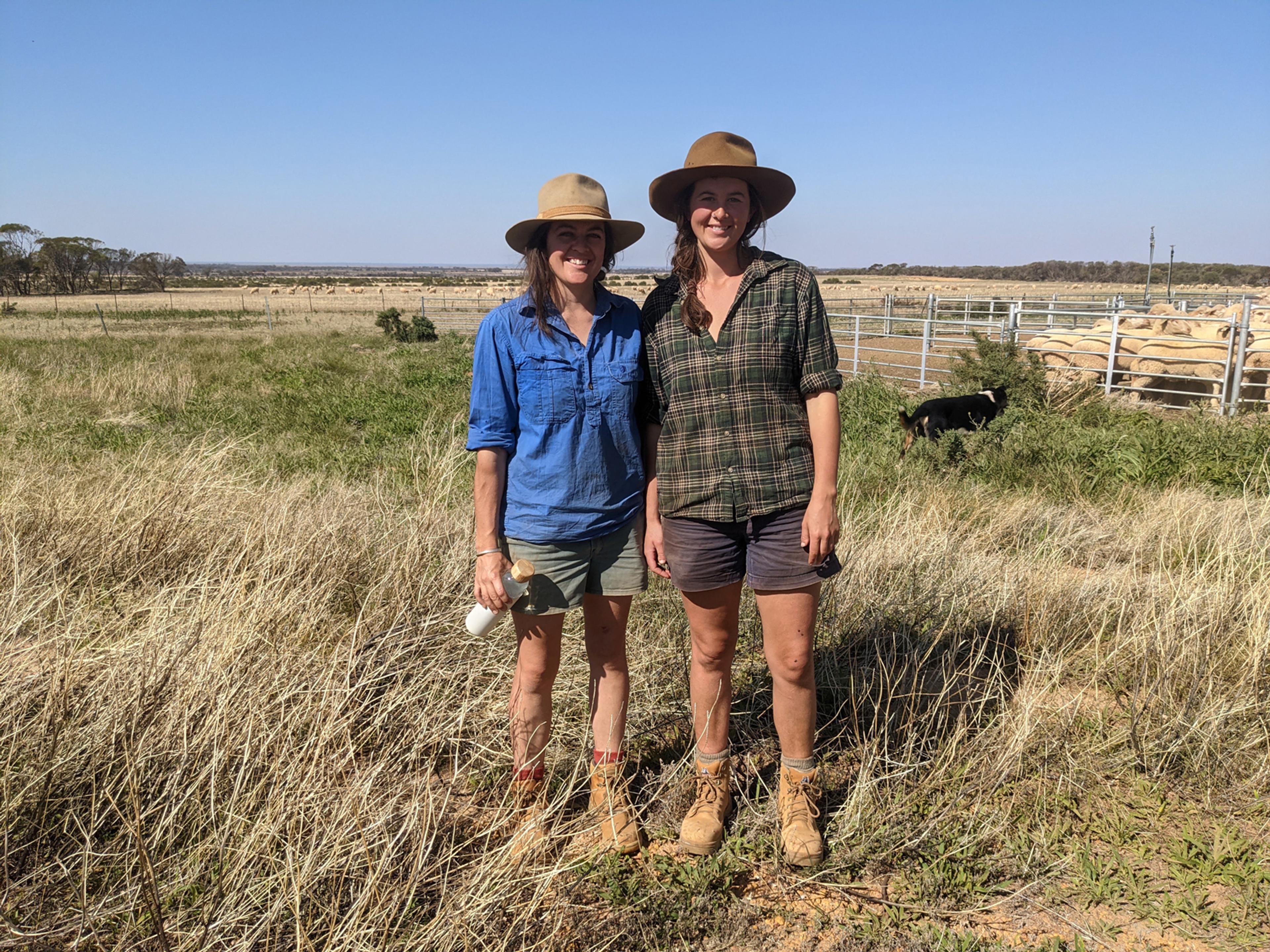Two people standing, smiling at the camera. They are both wearing flannelette shirts, shorts and wide brimmed hats. They are standing in a paddock with blue sky behind them.