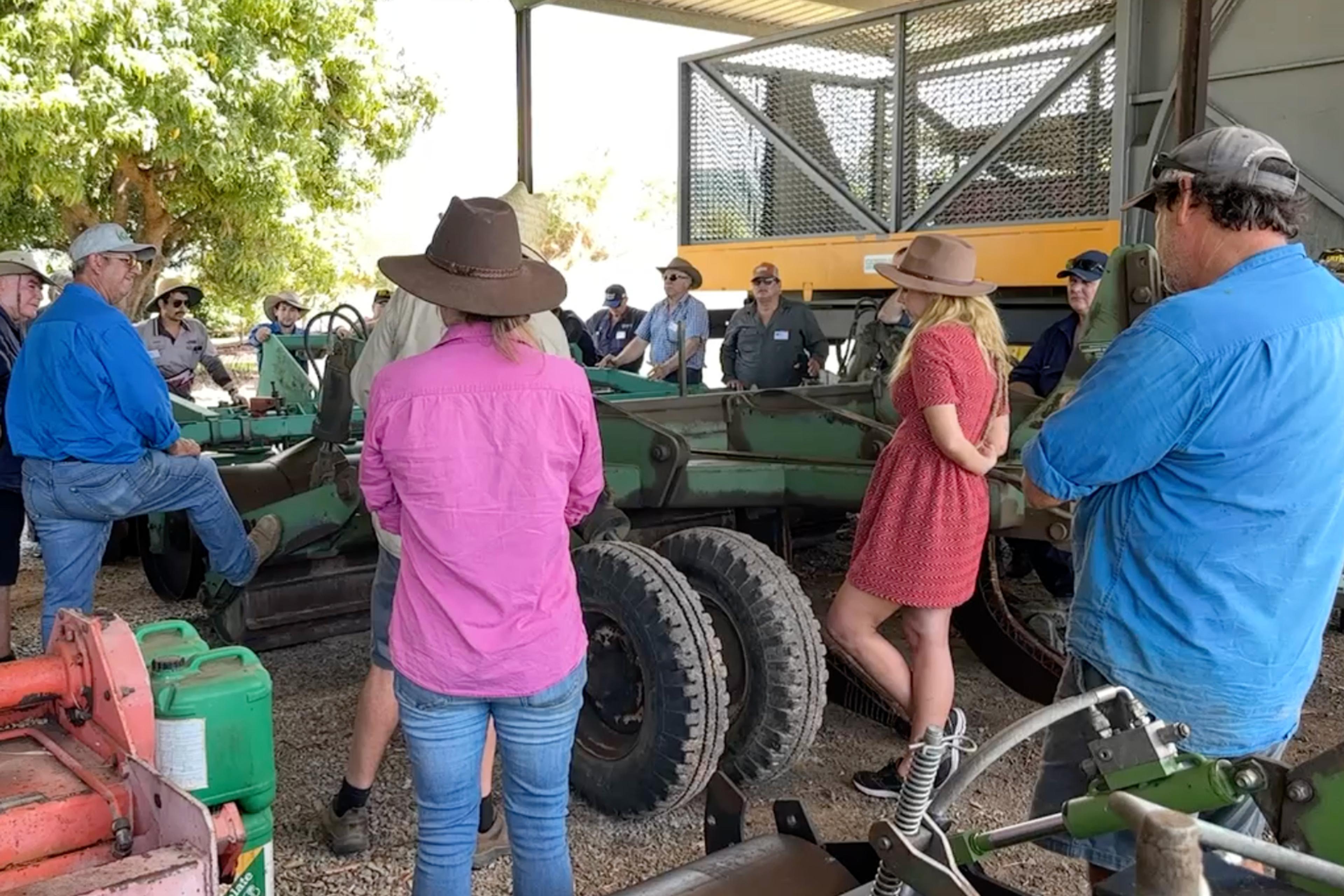 A group of people standing around a piece of machinery in an outdoor shed watching a person speaking.