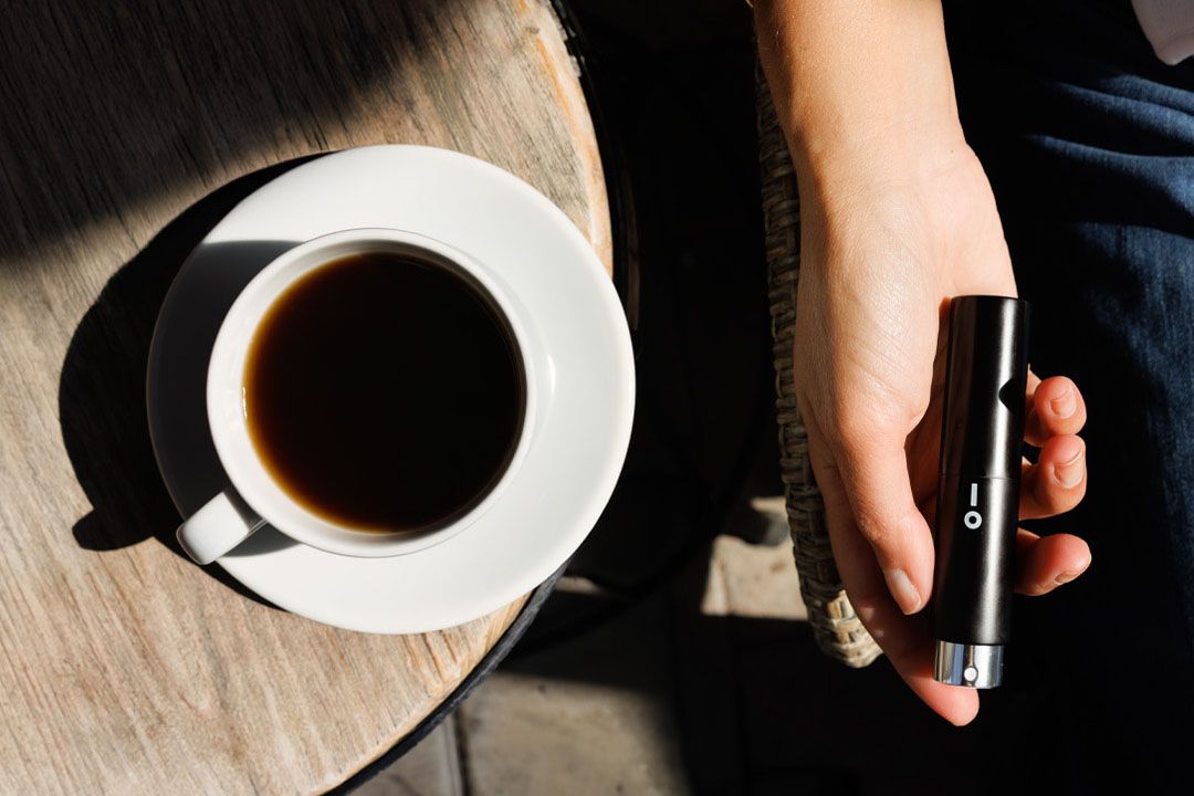 An overhead view of a café table with a coffee cup and a hand holding an 8 ml perfume bottle.