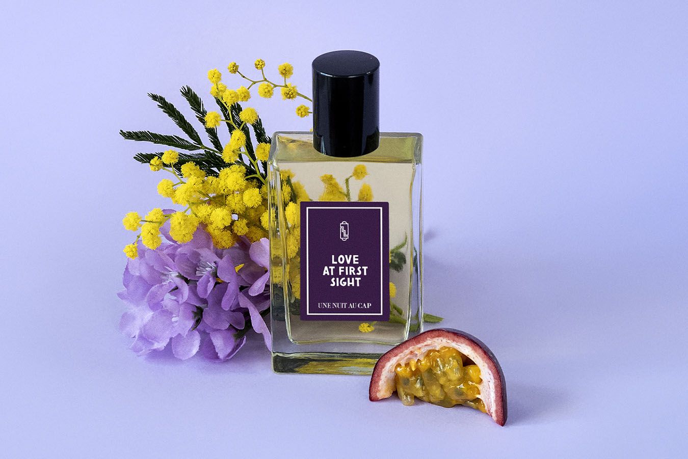 Une Nuit Nomade-parfymen Love At First Sight mot en ljuslila bakgrund, omgiven av mimosa, syréner och passionsfrukt