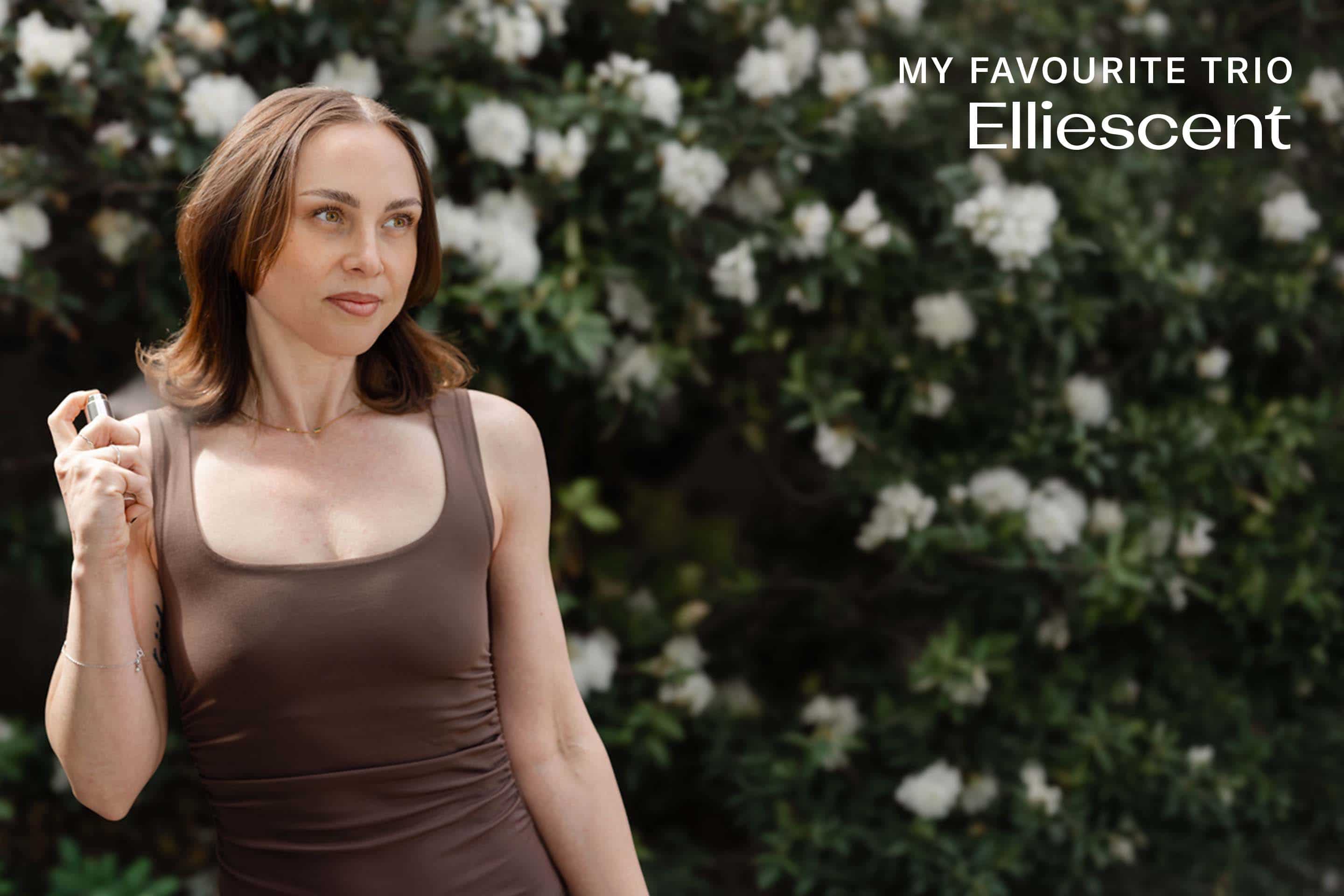 Ellie standing in front of a bush with white flowers, holding a silver colored perfume case. She has shoulder length brown hair, a brown top and is looking to the side.