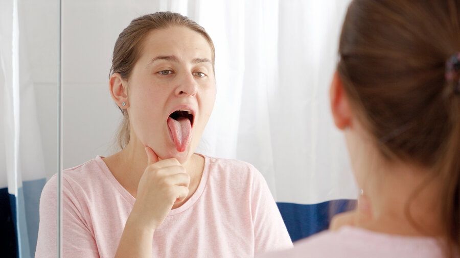 Young woman checking her teeth and tongue at mirror in bathroom. Learn about the oral microbiome.
