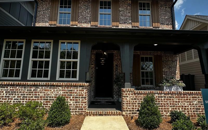 Front exterior of a two-story new construction home with antique brick facade, dark charcoal siding accents, white-trimmed windows, covered front porch with arched entry, dormer windows, and fresh landscaping with boxwood shrubs and pine straw
