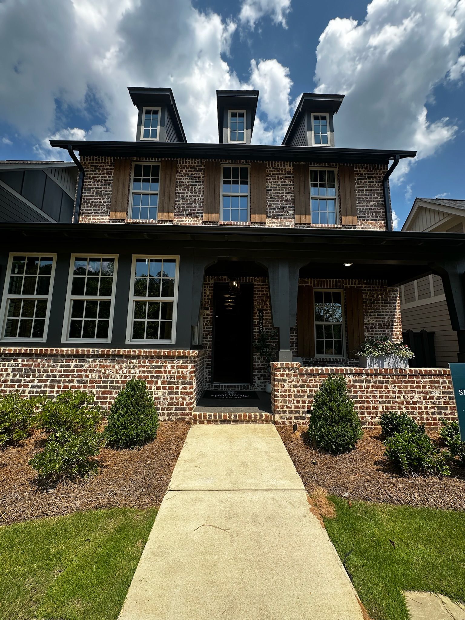 Front exterior of a two-story new construction home with antique brick facade, dark charcoal siding accents, white-trimmed windows, covered front porch with arched entry, dormer windows, and fresh landscaping with boxwood shrubs and pine straw