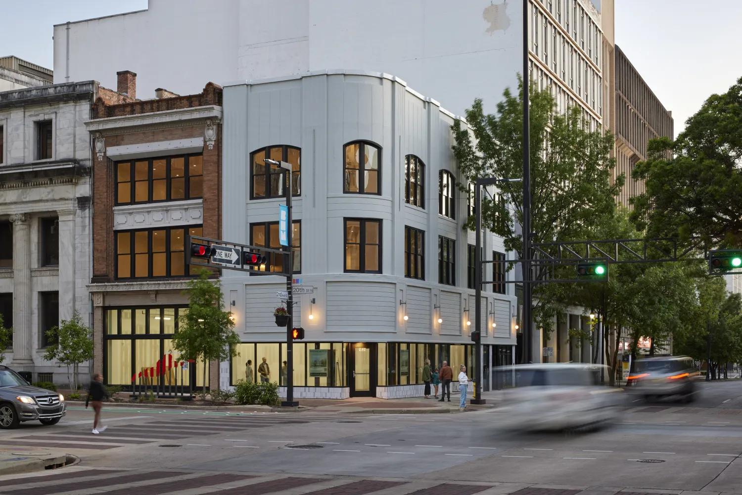Evening street scene in downtown Birmingham at 20th Street North, with a curved modern-renovated building glowing with warm light, pedestrians on the sidewalk, and cars in motion