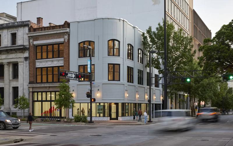 Evening street scene in downtown Birmingham at 20th Street North, with a curved modern-renovated building glowing with warm light, pedestrians on the sidewalk, and cars in motion