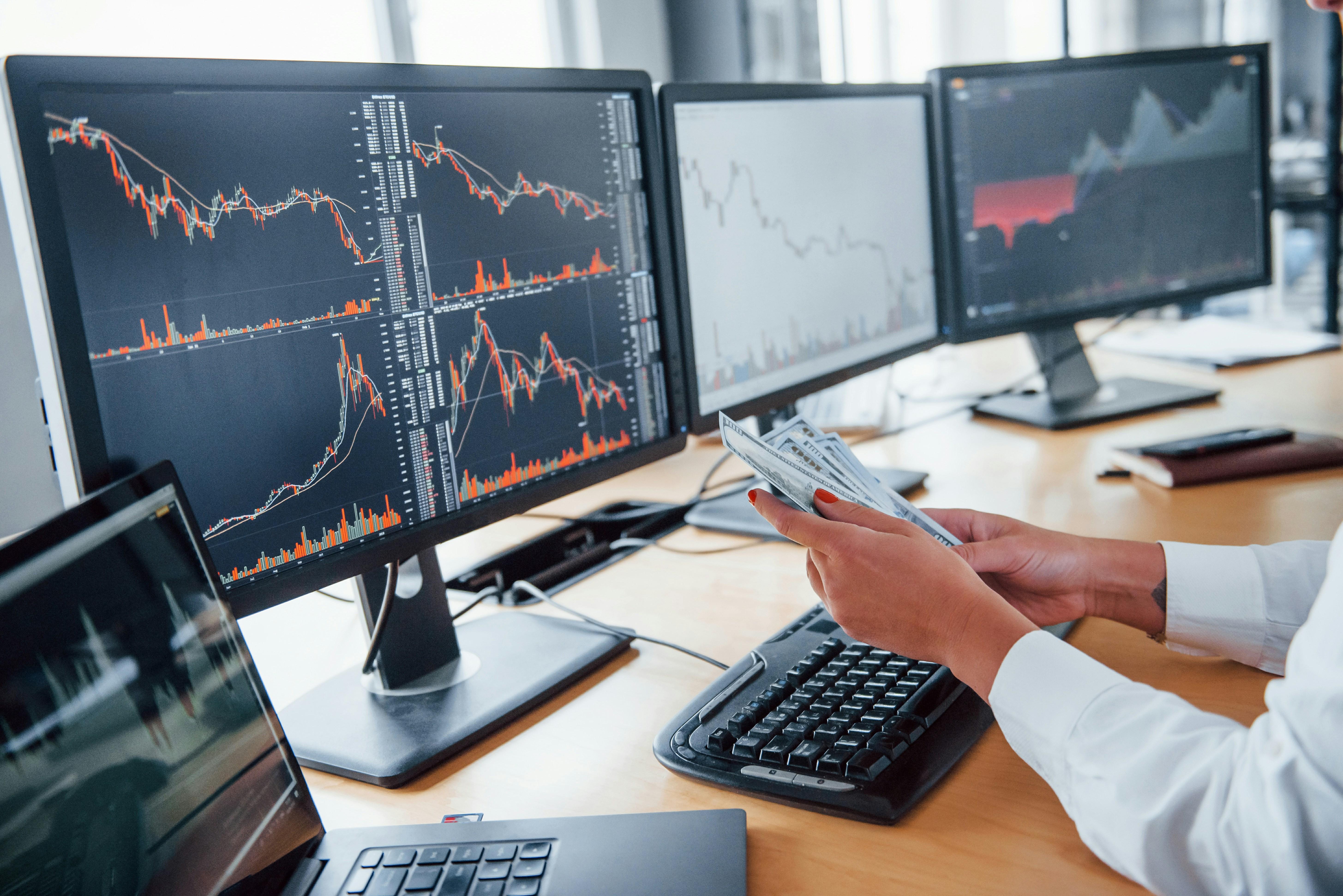 Person counting US dollar bills at a desk with multiple monitors displaying financial charts and stock market data.