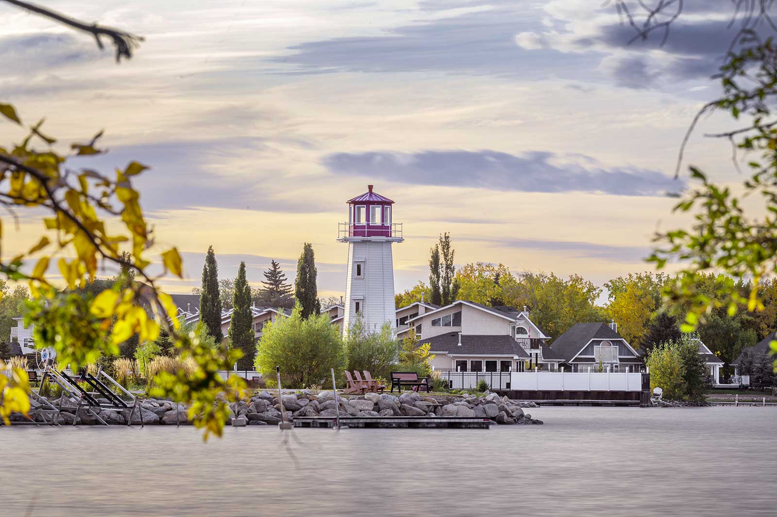 A red and white lighthouse by a lake, houses, trees.