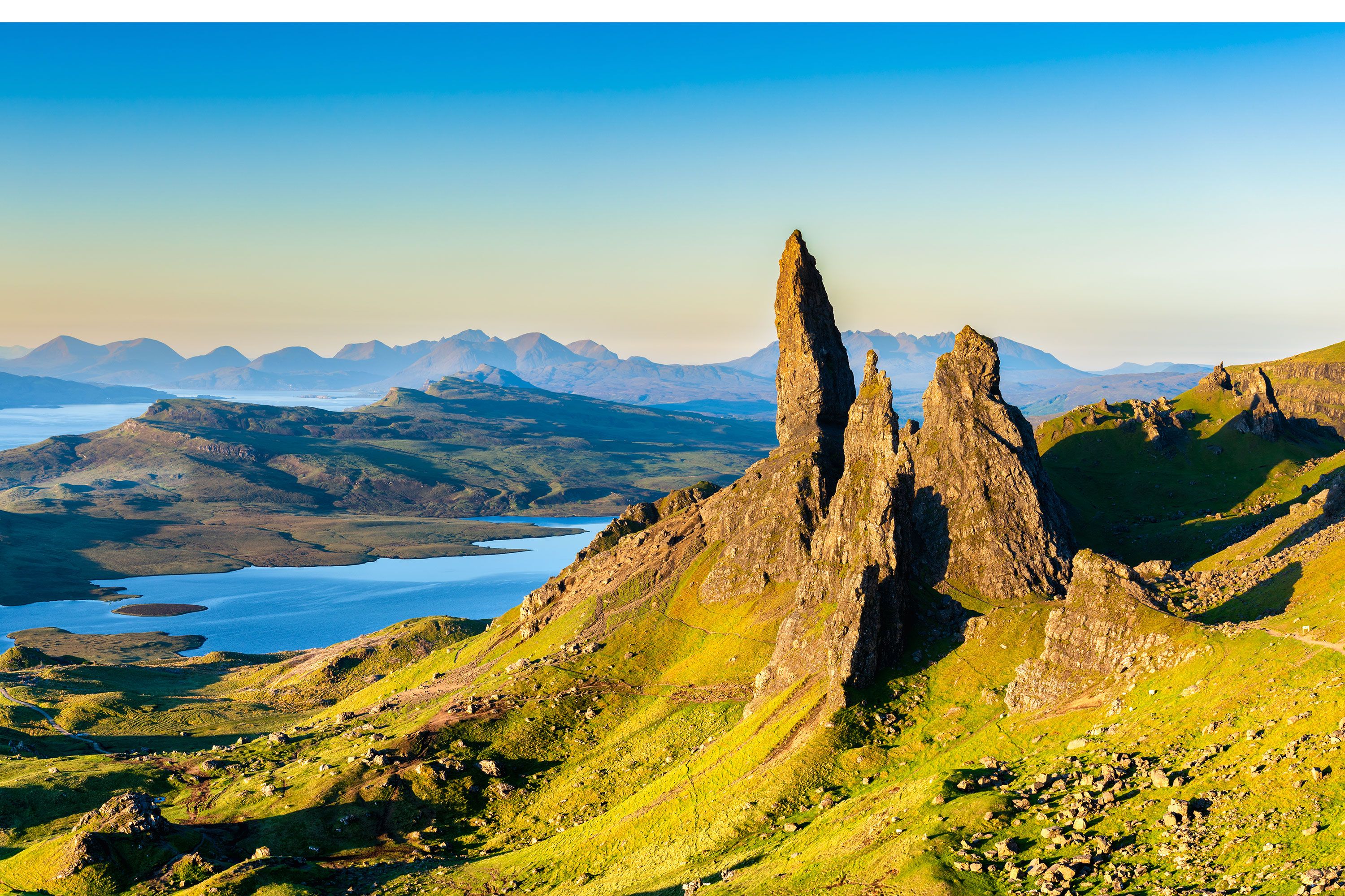 A large pointed rock formation in morning sunlight looking over a coastline and mountains.