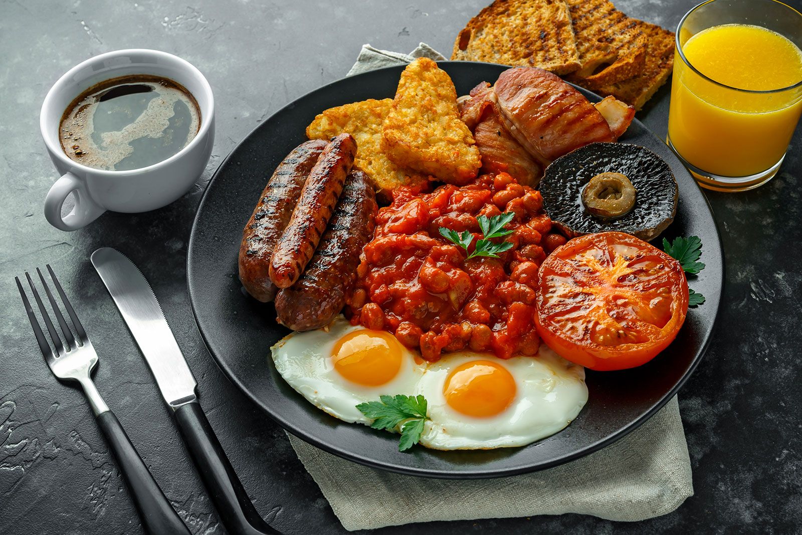 Full English breakfast with bacon, sausage, fried egg, baked beans, hash browns and mushrooms in black plate.