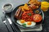 Full English breakfast with bacon, sausage, fried egg, baked beans, hash browns and mushrooms in black plate.