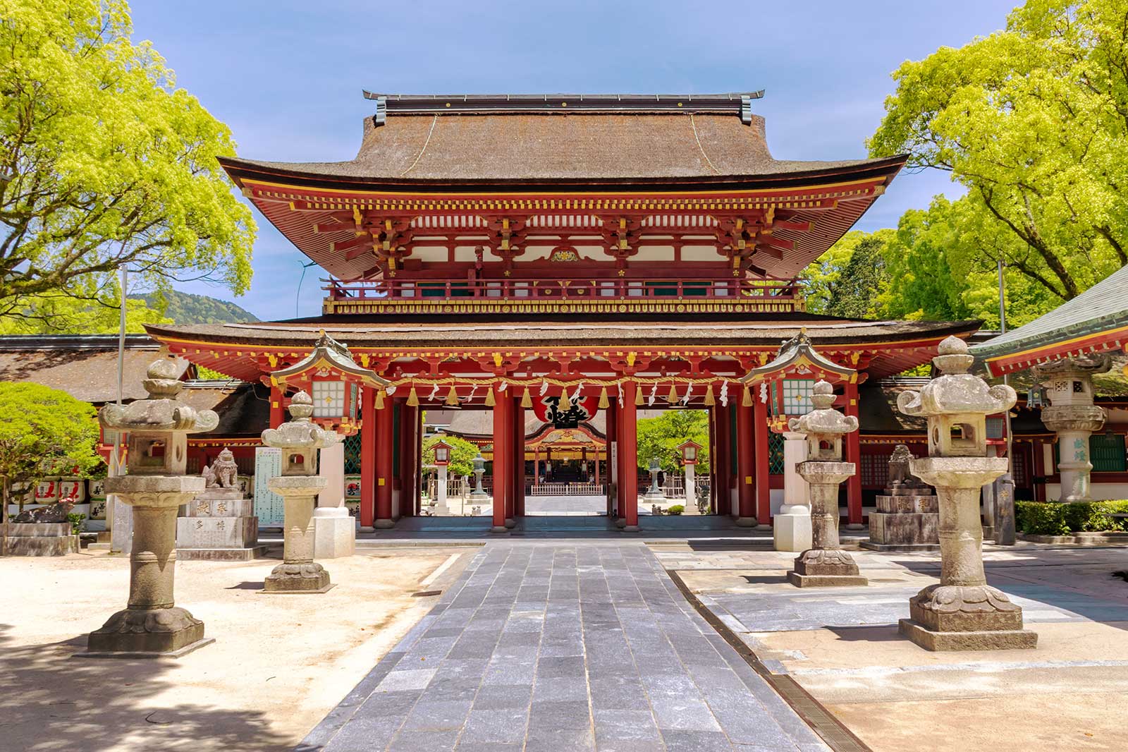 A red temple gate on a stone paved square, surrounded by green trees.