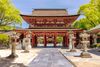 A red temple gate on a stone paved square, surrounded by green trees.
