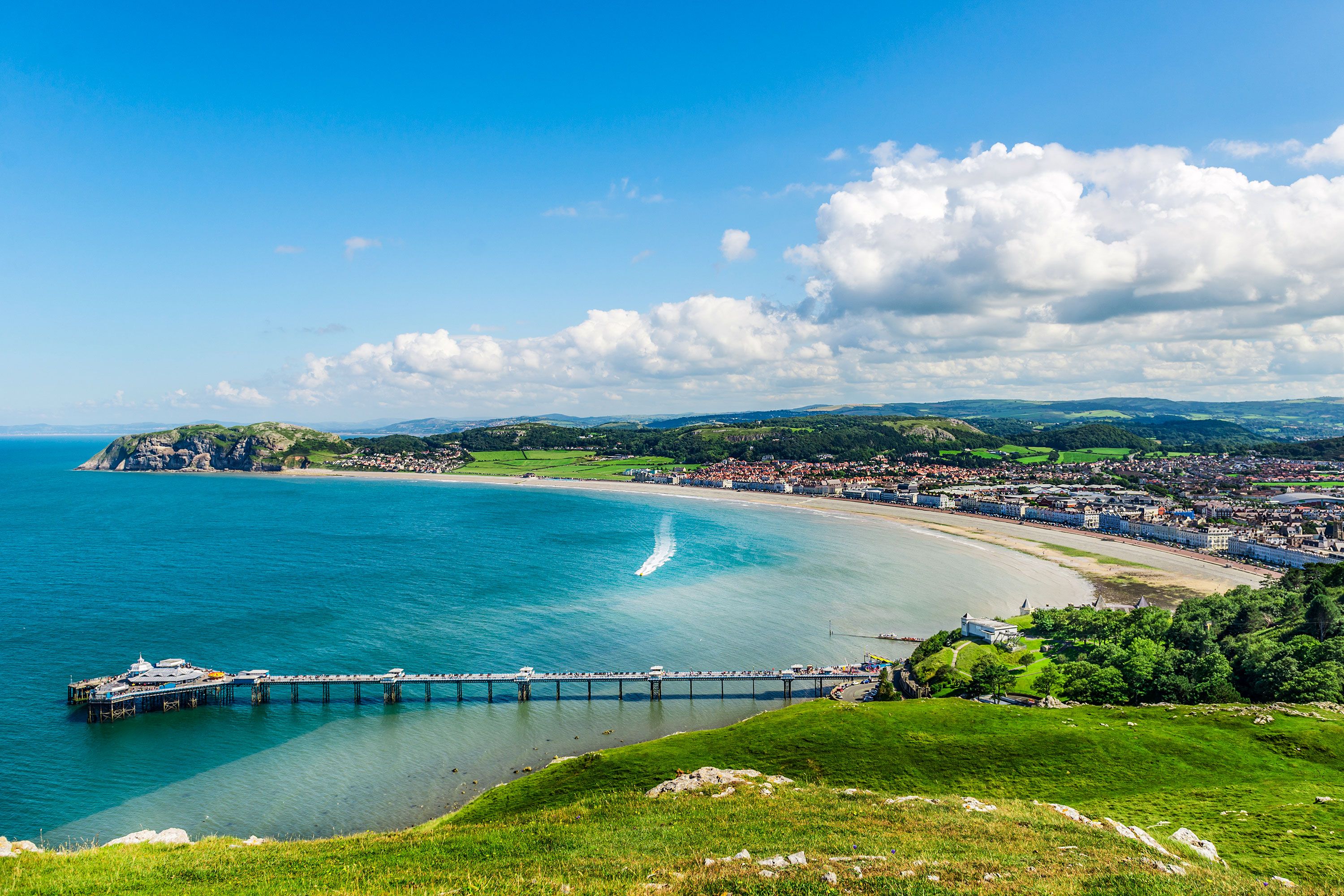 The Llandudno Sea Front in North Wales with a long pier and pavilion with a beach and town and rolling green hills in the distance.