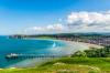 The Llandudno Sea Front in North Wales with a long pier and pavilion with a beach and town and rolling green hills in the distance.