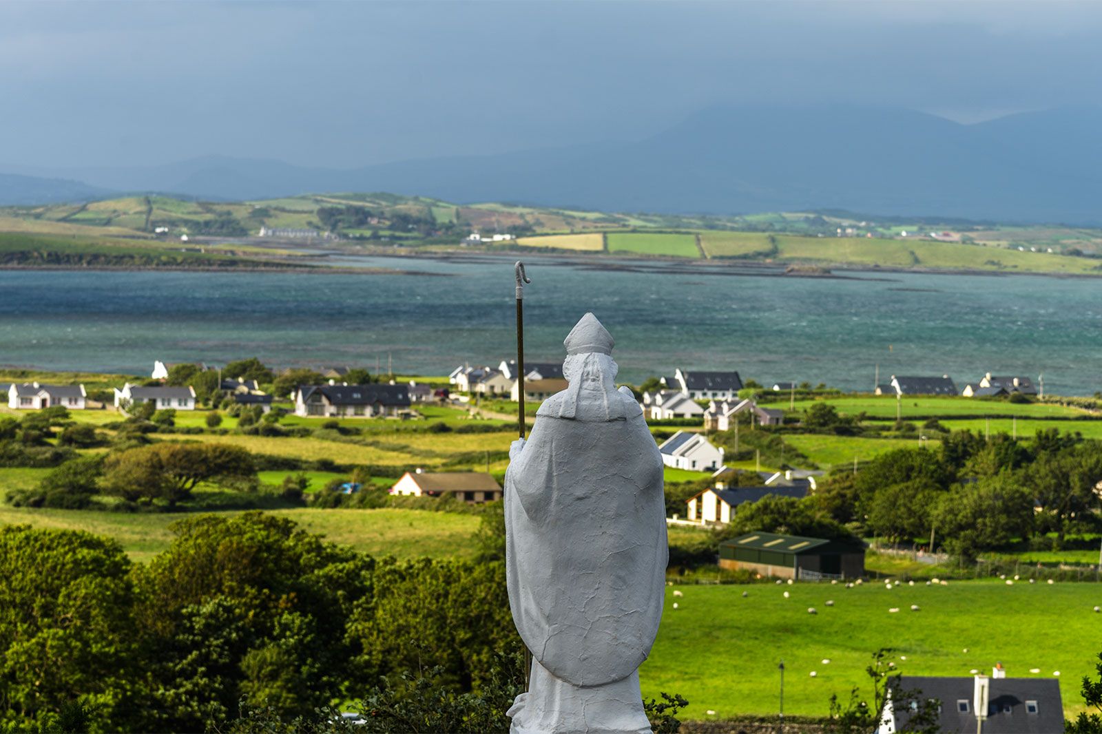 A statue overlooks green fields and mountains.