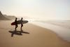 Two surfers with surfboards walk on the beach.