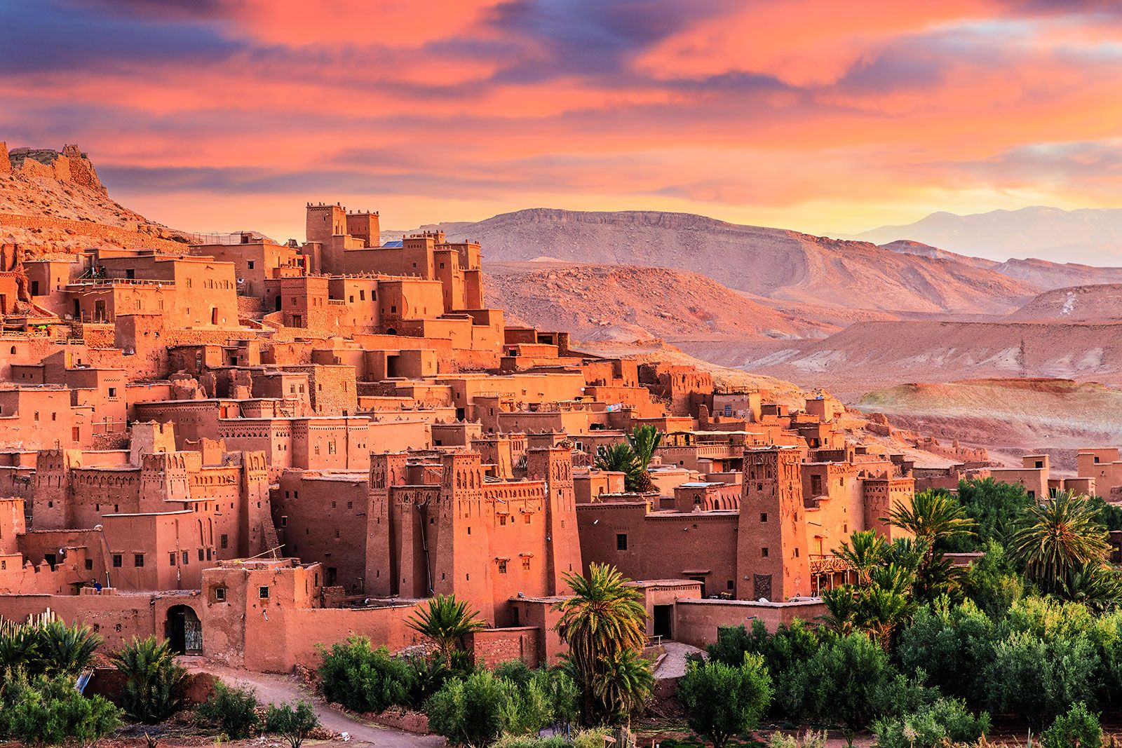 A multi-level sandstone town sits on a cliffside surrounded by greenery and mountains under a red-purple sky