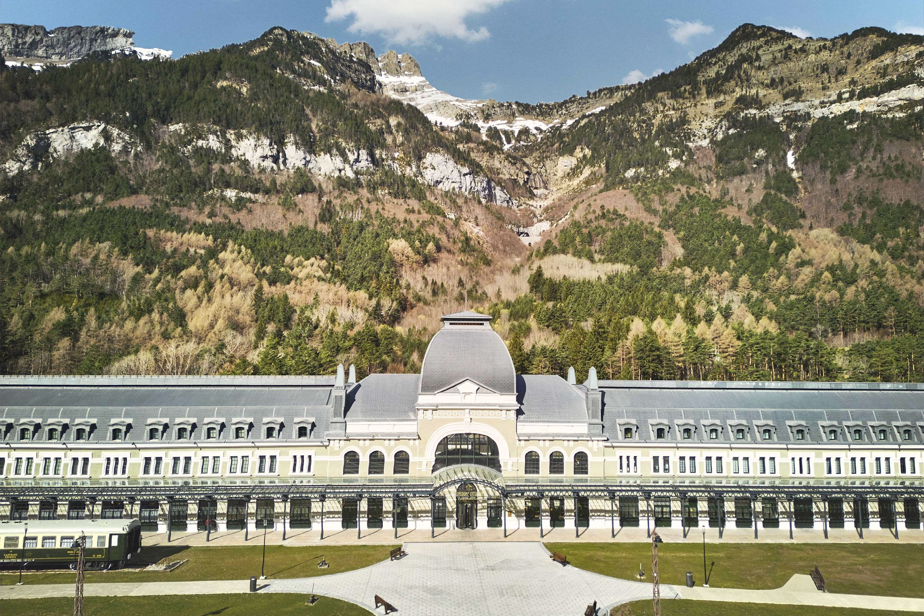 The exterior of a former train station turned into a hotel with mountains behind it.