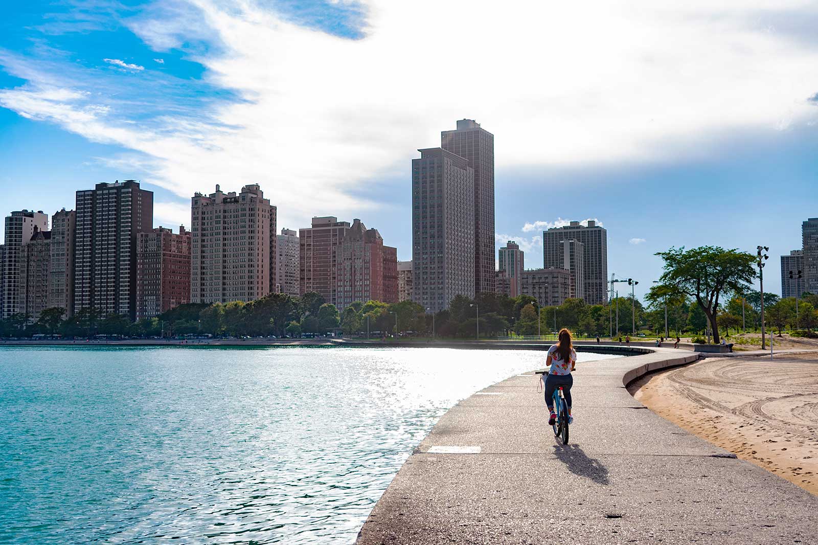  Woman on a bicycle pedaling by a beach with the Chicago skyline.