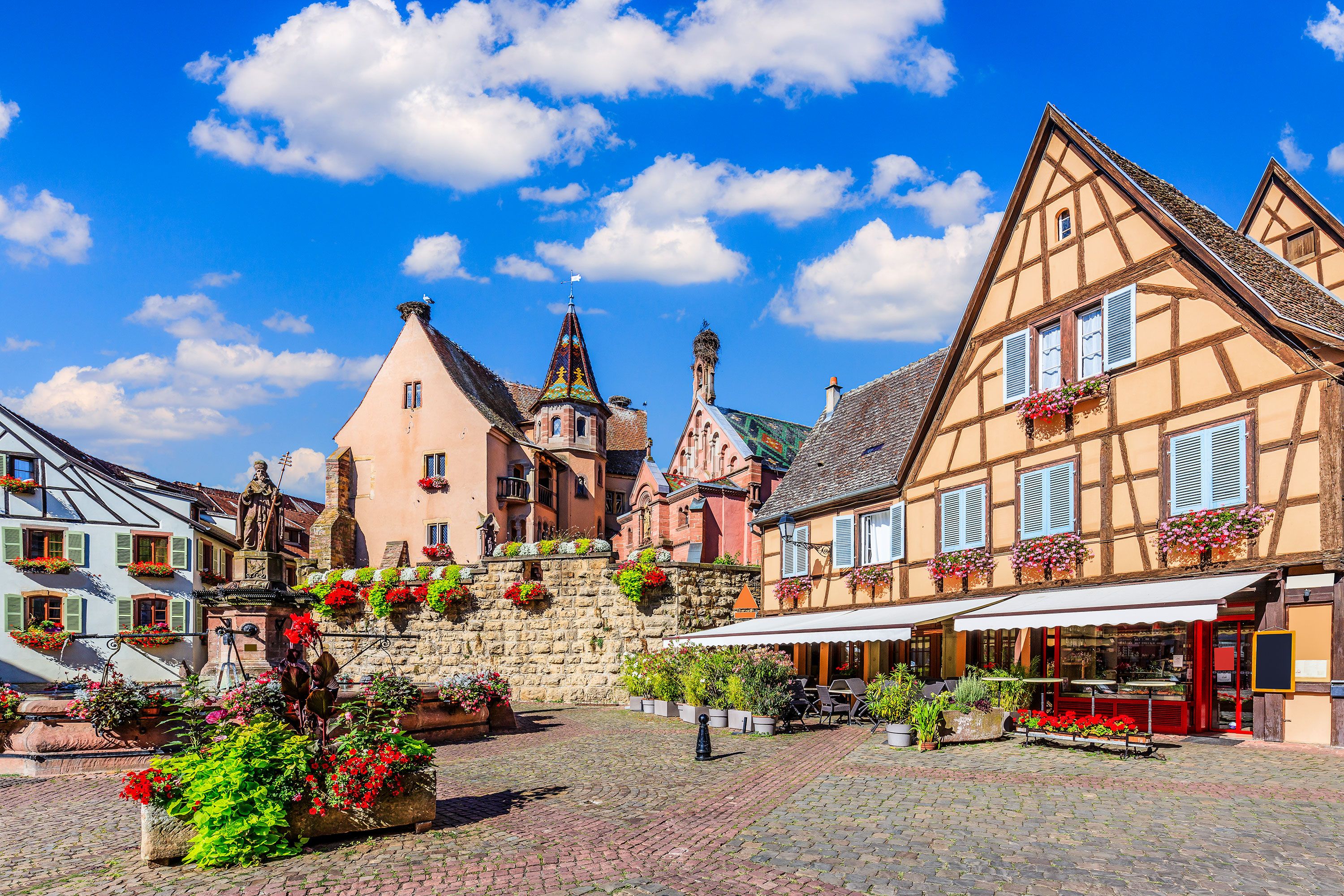 Colorful Alsatian buildings surround a cobblestone plaza decked with planters and flowers.