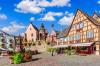 Colorful Alsatian buildings surround a cobblestone plaza decked with planters and flowers.