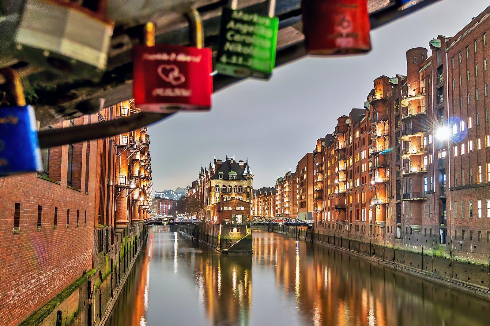 Lights from a building is reflected in water on a canal with locks connected to a bridge in the foreground.