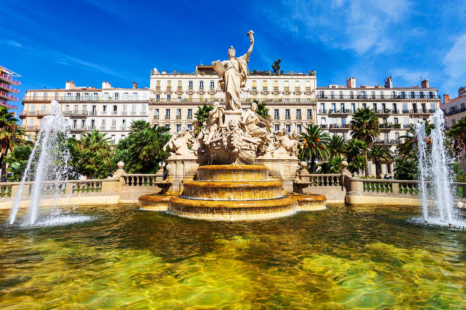 A fountain with a statue in the centre, buildings in the background.