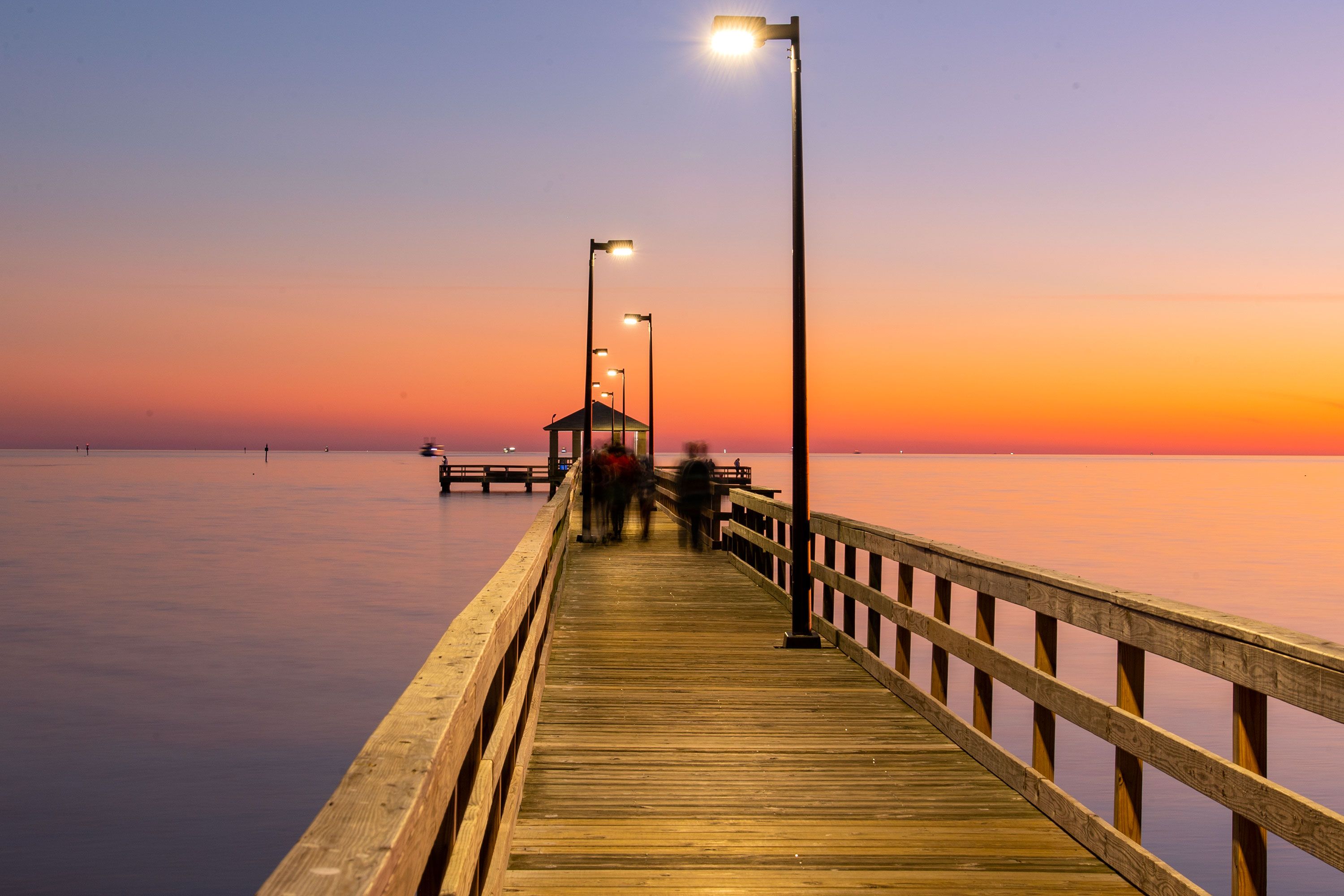 A lit wooden pier stretches over calm water at sunset, with blurred figures walking towards a gazebo.