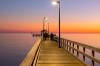A lit wooden pier stretches over calm water at sunset, with blurred figures walking towards a gazebo.