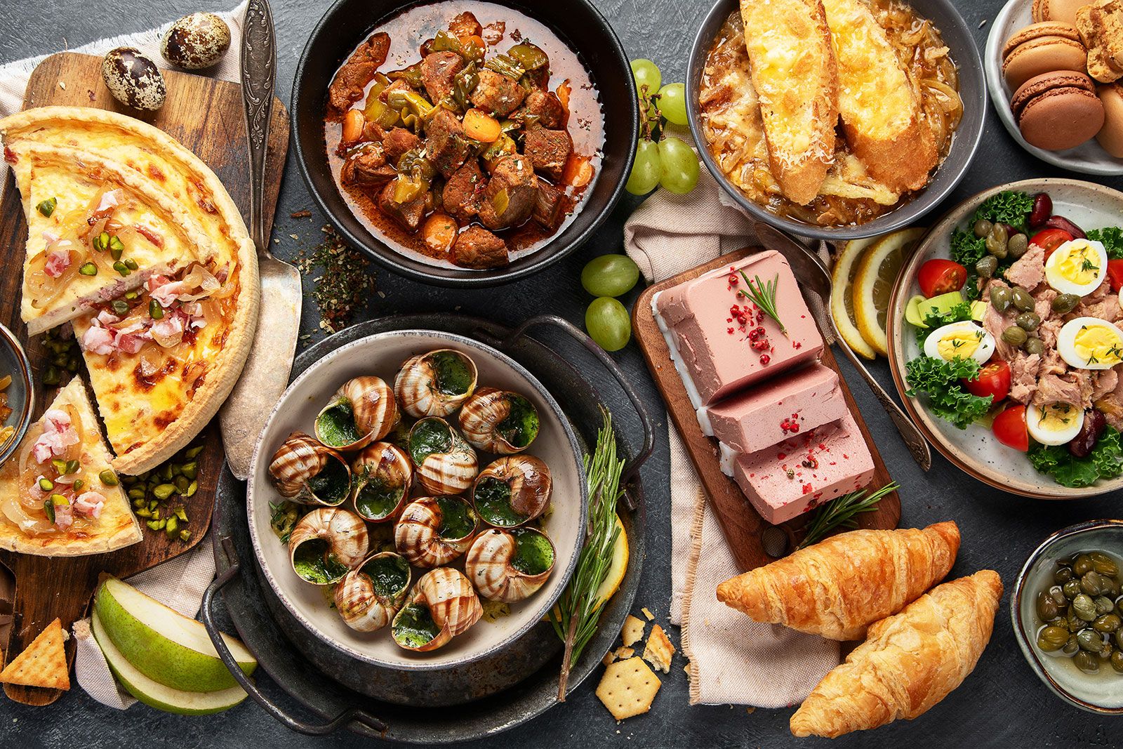 A table with plate containing french food -mussel and meat dishes, ratatouille, snails, croissants, desserts, fresh pastries and cheese.