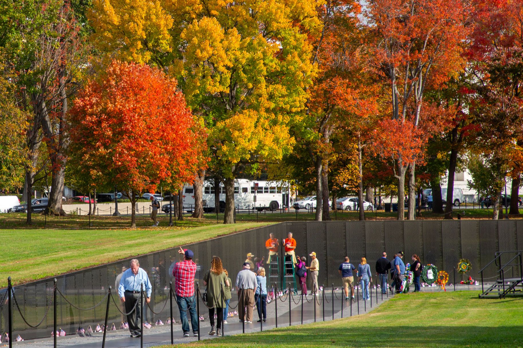 People walk along a memorial with fall colored trees in the background.