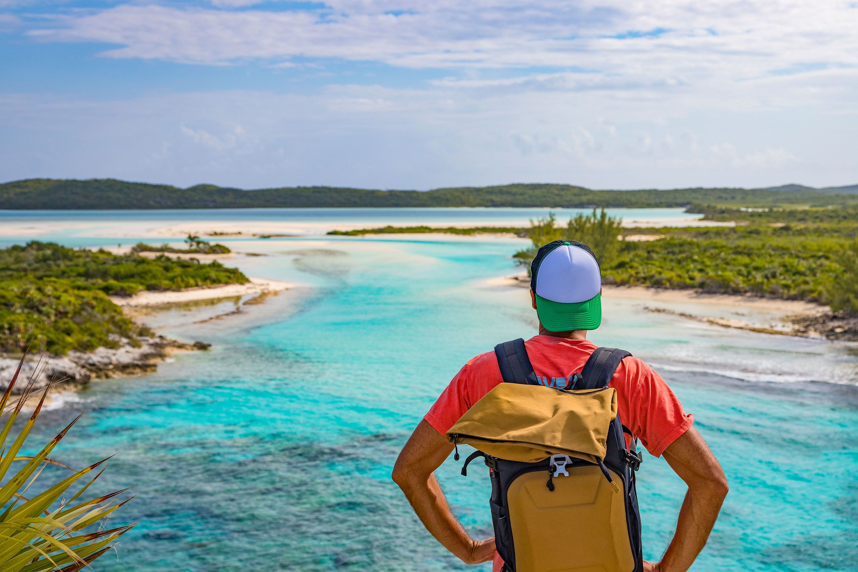 Young man in orange shirt with a yellow backpack in the foreground faces the light blue water with green land and beaches on the right and left that lead out to open sea.
