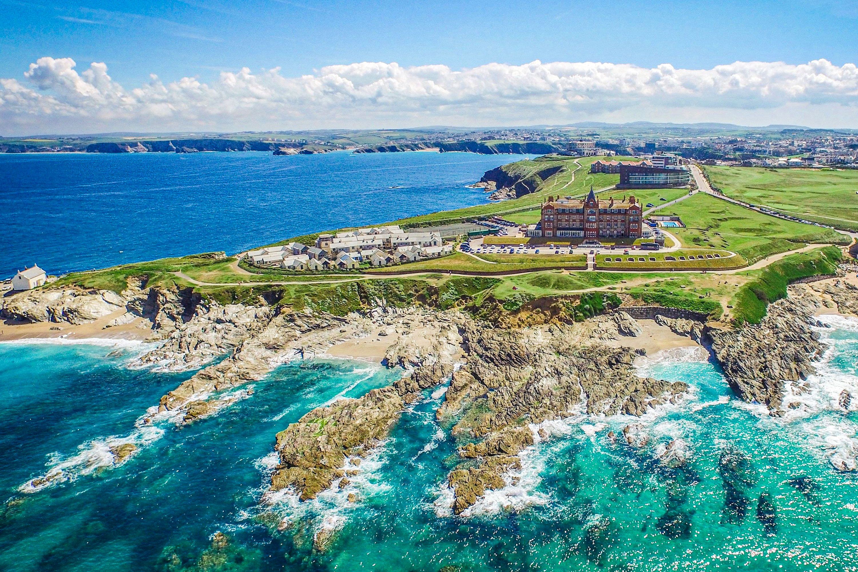 Aerial view of rocky point jutting out into the water with green grass and sandy coves with multiple smaller structures and one larger castle-like building with a town, hills, and puffy clouds in background.
