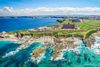 Aerial view of rocky point jutting out into the water with green grass and sandy coves with multiple smaller structures and one larger castle-like building with a town, hills, and puffy clouds in background.