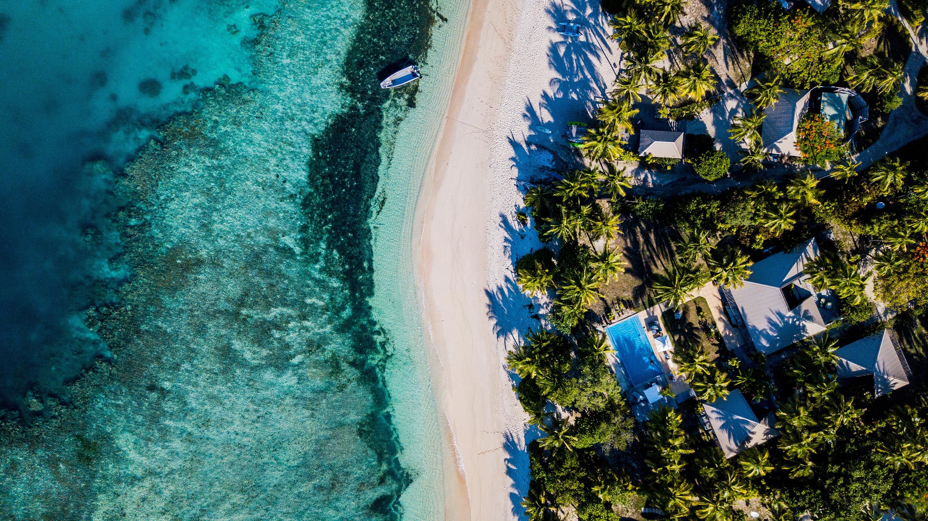 An aerial view of crystal-clear waters crashing onto a beach and houses among palm trees in Fiji.