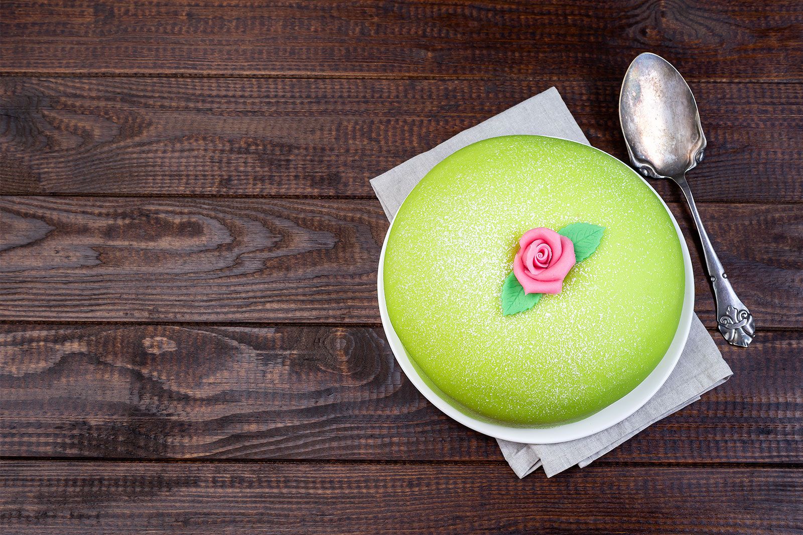 A green cake with a pink rose on wooden table and a fork.