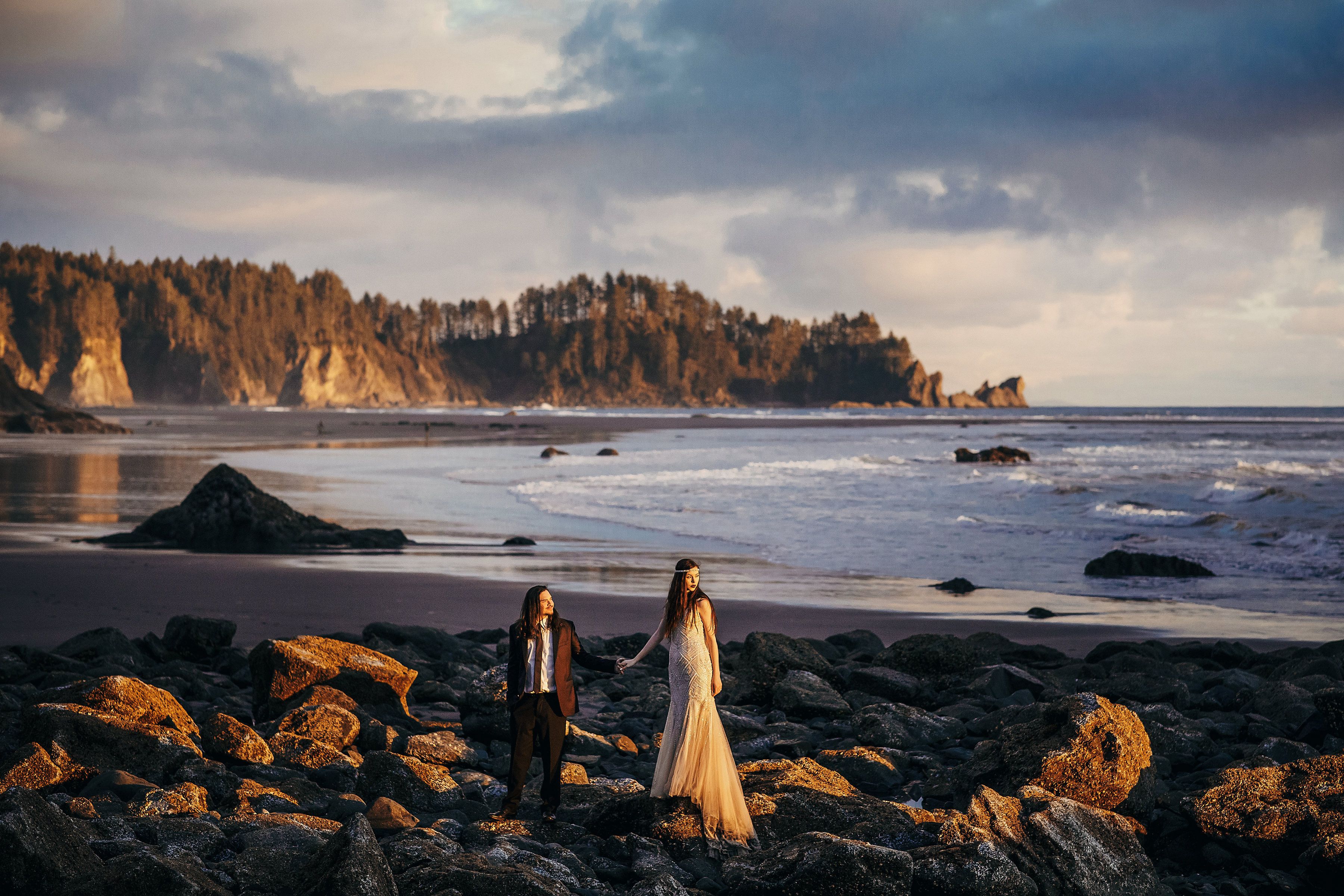  A bride and groom holding hands and standing over rocks near beach and looking at the sunset.