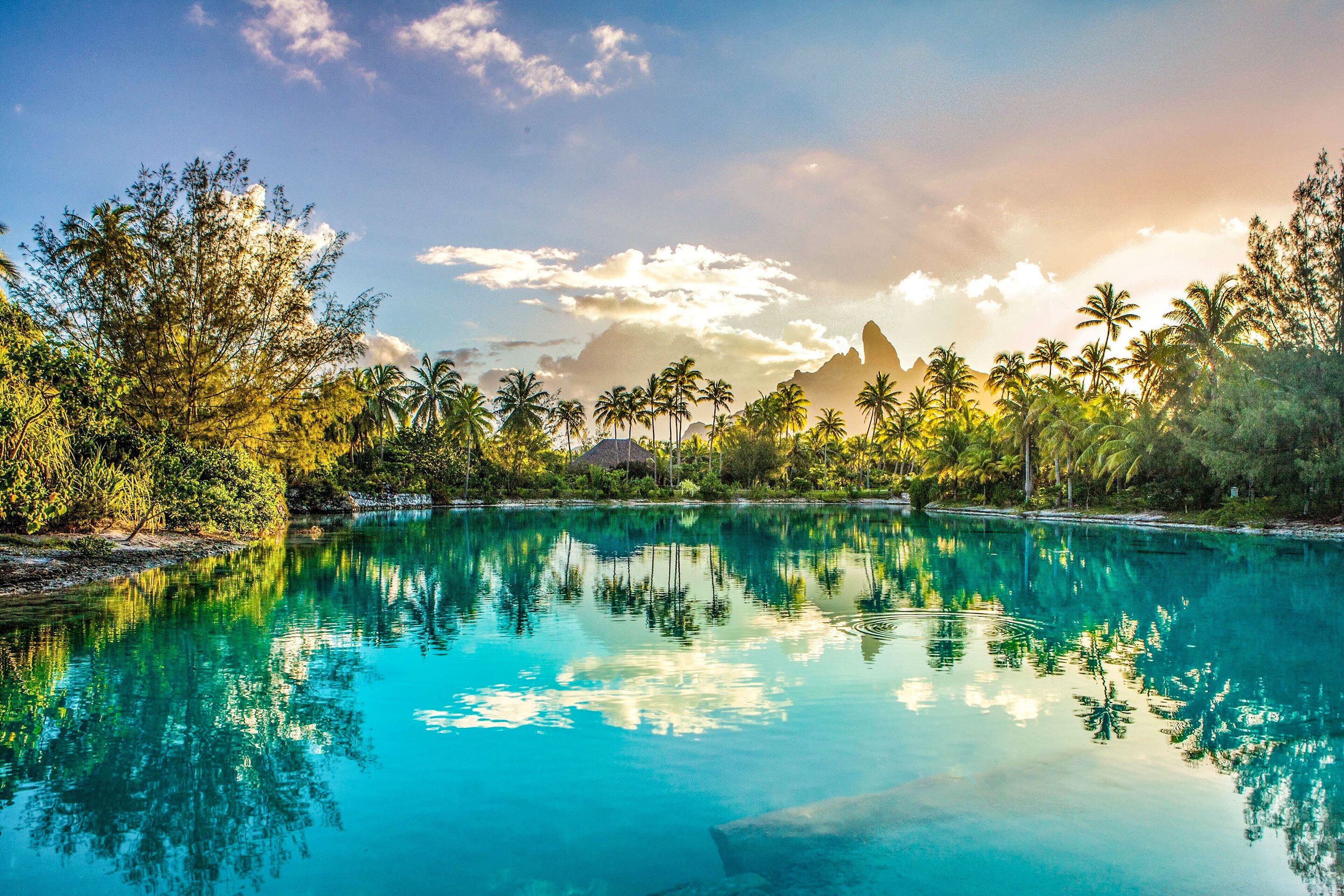 A blue lagoon with thin sandy shore is ringed by palm trees and vegetation with a mountain in the background with the sun shining through clouds.