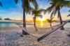 Hammock hangs between two palm trees during a sunset on a sandy beach.