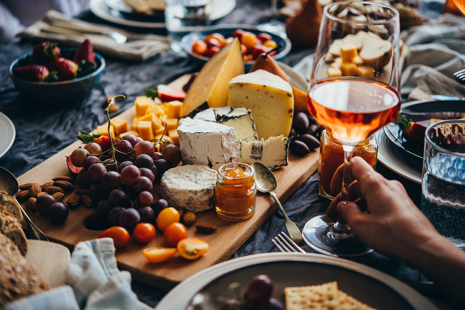 A closeup of hand holding a wine glass with different types of cheese and fruits kept on the table.
