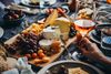 A closeup of hand holding a wine glass with different types of cheese and fruits kept on the table.