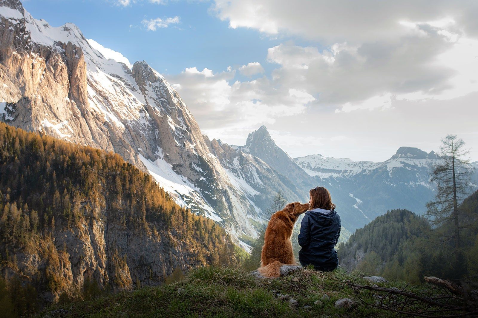 A dog with its owner sitting on the ground, mountains in the background.