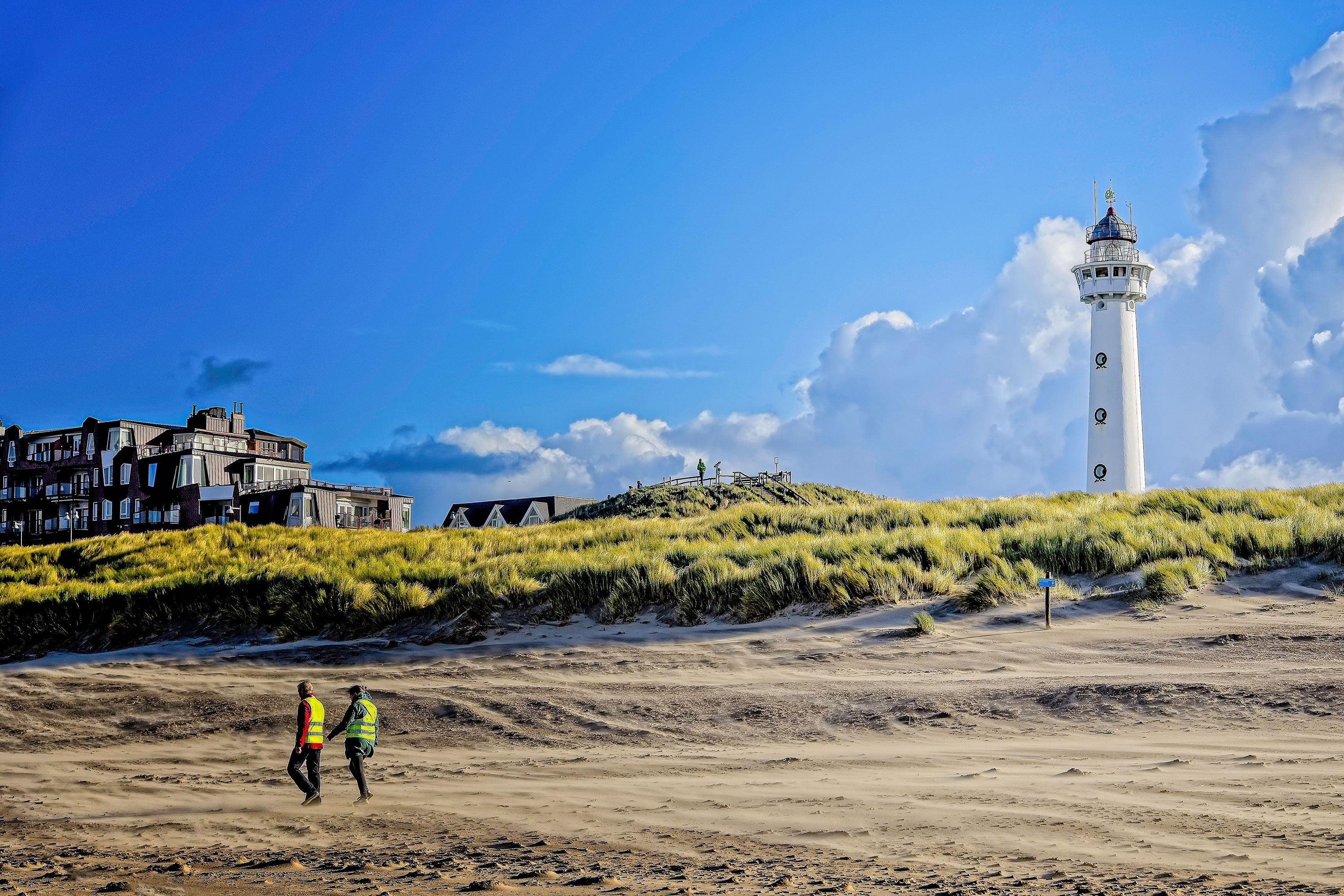 A couple stroll on Egmond aan Zee in Leuchtturm with dunes and buildings and a lighthouse in front of dramatic white clouds in the background.