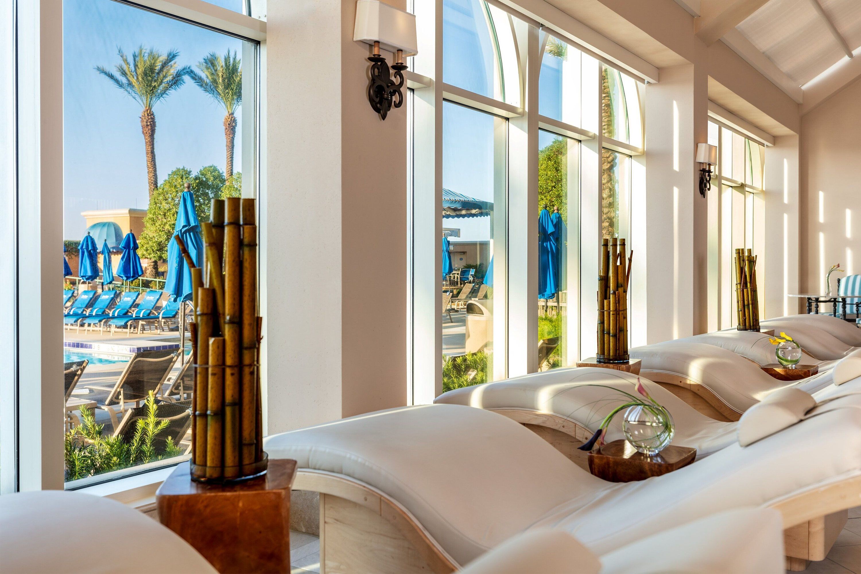 White cushioned spa loungers line a sunlit relaxation room overlooking a pool with blue umbrellas and palm trees.