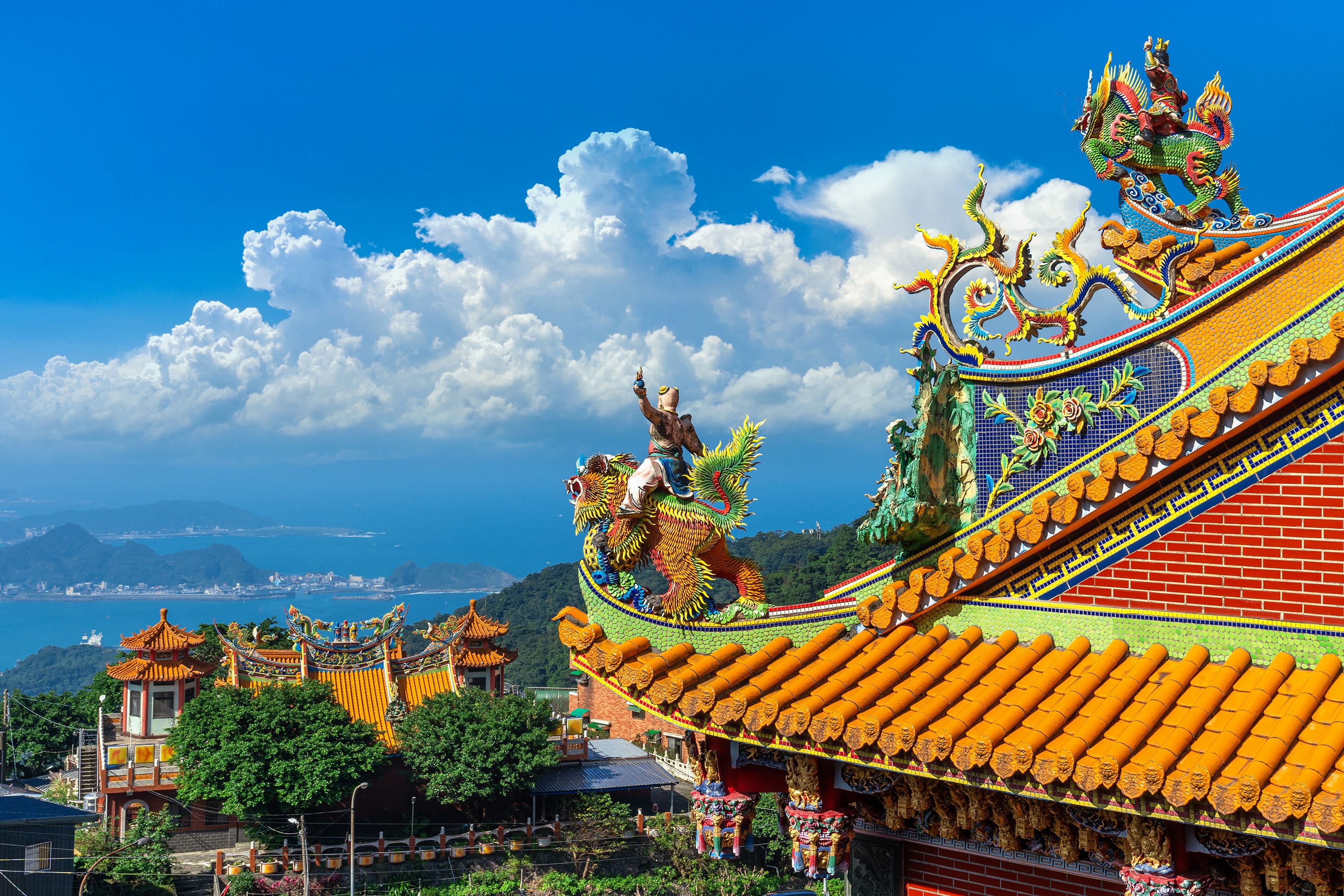 Aerial view looking at a roof of a temple with person riding a dragon and another temple and sea off in the distance.